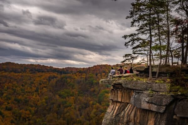 Hikers sitting at Diamond Point Overlook in New River Gorge, with autumn foliage and a dramatic cloudy sky in the background.